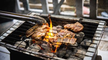 Piknik partisinde sıcak dumanlı kömür ızgarasında pişirilmiş metal maşayla sosla marine edilmiş çiğ kuzu eti tutan eli yakından izleyin. Önplanda seçmeli odak.