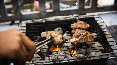 Piknik partisinde sıcak dumanlı kömür ızgarasında pişirilmiş metal maşayla sosla marine edilmiş çiğ kuzu eti tutan eli yakından izleyin. Önplanda seçmeli odak.