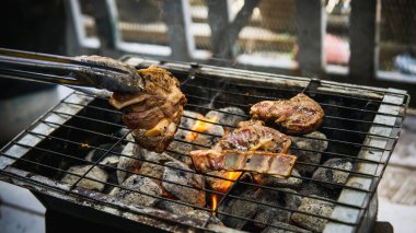 Piknik partisinde sıcak dumanlı kömür ızgarasında pişirilmiş metal maşayla sosla marine edilmiş çiğ kuzu eti tutan eli yakından izleyin. Önplanda seçmeli odak.