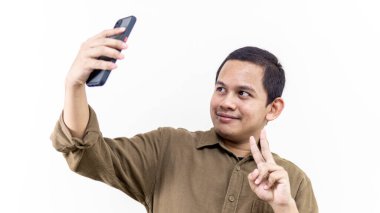 Genç Asyalı Malezyalı adam gülümseyen bir selfie portresi çekiyor ve izole beyaz arka planda akıllı telefon kullanarak barış işaretini parmağıyla gösteriyor..
