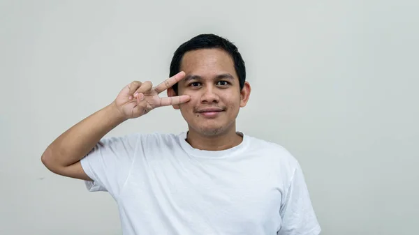 一幅快乐的亚裔马来人画像 身穿白色T恤 面带微笑 用手指对着相机 在孤立的白色背景上签着和平字 心情又好又开心 — 图库照片