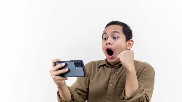 一位身穿棕色衬衫的年轻的亚裔马来人满脸喜色地看着智能手机屏幕上孤立的白色背景 庆祝和享受这个好消息 — 图库照片