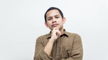 Asyalı genç Malezyalı 'nın kafası karışmış ve düşünceli yüz ifadesi. Eli çenesinde, gömleği ise izole edilmiş beyaz bir arka planı var. Düşünme konsepti.