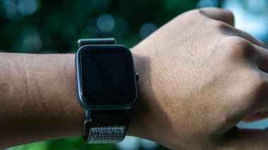 Yakından bakınca bileğinde siyah dokunmatik ekranı olan akıllı bir adam görüyorum. O, saat şeklinde takılabilir bir bilgisayardır. Verileri akıllı telefona senkronize edebiliyor. Nesneye seçici odaklanma.