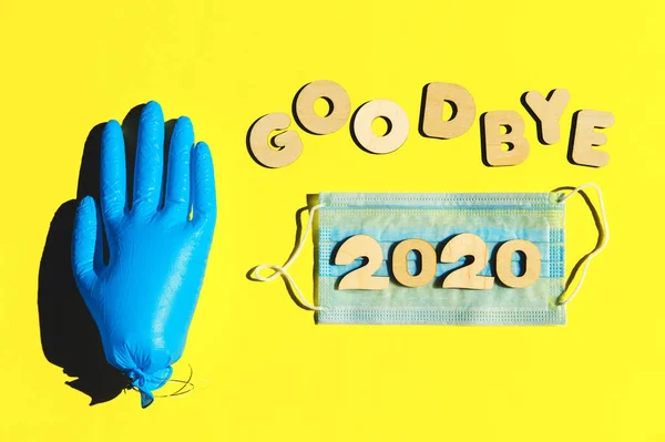 Ord GOODBYE 2020 från träbokstäver på gul bakgrund, uppblåst medicinsk handske och ansiktsvatten. Koncept för 2020 och epidemi. — Stockfoto