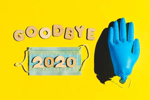 Λέξεις GOODBYE 2020 από ξύλινα γράμματα σε κίτρινο φόντο, φουσκωμένο ιατρικό γάντι και μάσκα προσώπου. 2020 και έννοια της επιδημίας. — Φωτογραφία Αρχείου