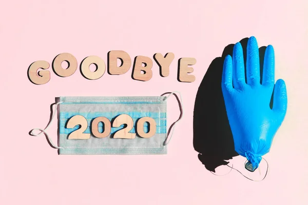 Ord GOODBYE 2020 från träbokstäver på rosa bakgrund, uppblåst medicinsk handske och ansiktsvatten. Koncept för 2020 och epidemi. — Stockfoto