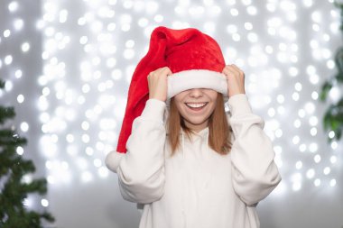 Genç bir kadın gözlerini kırmızı bir Noel Baba şapkasıyla kapatıyor ve gülüyor. Bokeh of Christmas çelenklerinin arkasında