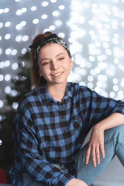 Young woman sits against the background of bokeh of Christmas garlands and a Christmas tree wearing casual clothes and smiles. Grainy film effect. — Photo