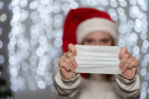 A young woman in a red Santa hat, pointing with her hands to a protective mask, stands against the background of bokeh of Christmas garlands. — Stock Photo, Image