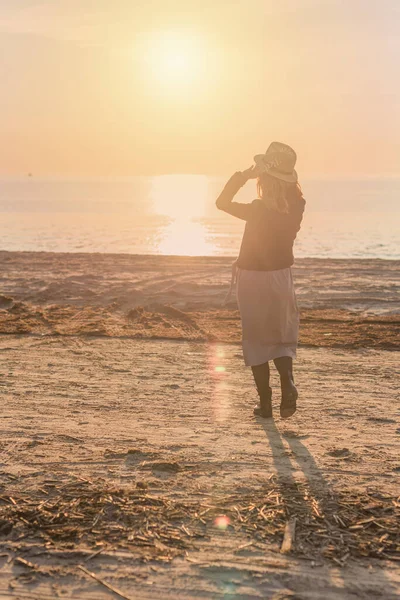 Эффект зернистой пленки на картинку. силуэт женщины в шляпе и платье, идущей вдоль пляжа на фоне заходящего солнца — стоковое фото