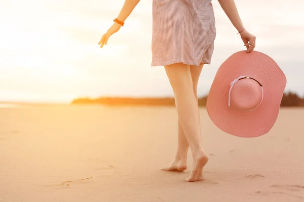 Zbliżenie żeńskich stóp spacerujących wzdłuż piaszczystej plaży, aby spotkać wschód słońca. Młoda kobieta w krótkiej sukience z kapeluszem w rękach spaceruje po plaży w słońcu. — Zdjęcie stockowe