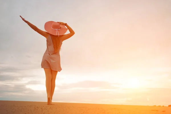 Силуэт романтичной и мечтательной молодой женщины, спускающейся по склону песчаной дюны с поднятыми руками. Женский силуэт в шляпе и короткое платье на фоне закатного неба. копировальное пространство — стоковое фото