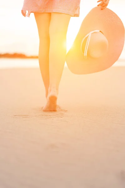Крупным планом женских ног, идущих вдоль песчаного пляжа, чтобы встретить восход солнца. молодая женщина в коротком платье с шляпой в руках идет по пляжу на солнце. — стоковое фото