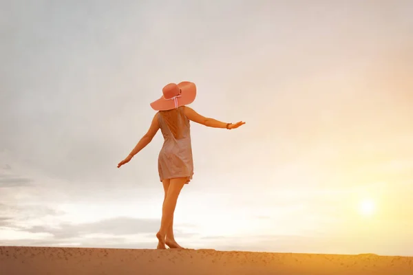 Силуэт романтичной и мечтательной молодой женщины, спускающейся по склону песчаной дюны с поднятыми руками. Женский силуэт в шляпе и короткое платье на фоне закатного неба. — стоковое фото