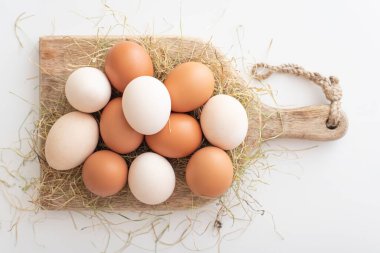 Tavuk yumurtaları tahta bir tahtanın üzerinde beyaz arka plandaki kuru otların içinde yatar. Paskalya yumurtalarını bayram için hazırlıyorum. Paskalya masasında açık ve kahverengi yumurtalar