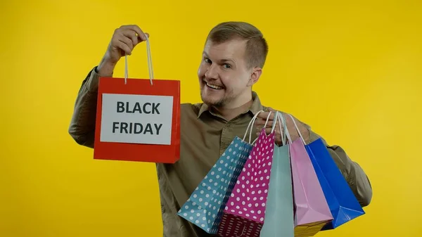 Hombre mostrando la inscripción Viernes Negro en bolsas de compras, sonriendo, satisfecho con las compras a precios bajos — Foto de Stock