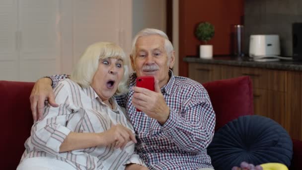 Abuela y abuelo haciendo videollamada en línea en el teléfono móvil en casa. Sonriendo pareja mayor — Vídeos de Stock