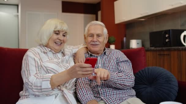 Szczęśliwa uśmiechnięta para seniorów z komórką w domu. Odpoczynek na kanapie w przytulnym salonie — Wideo stockowe