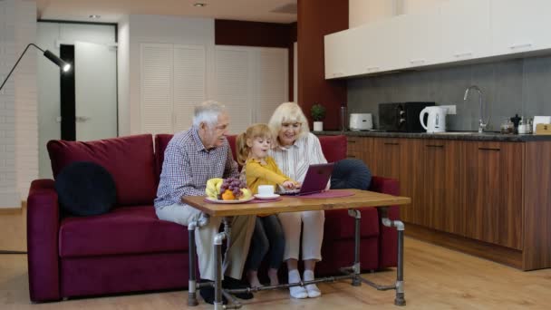 Dziadek i babcia siedzą w salonie i uczą małą wnuczkę za pomocą laptopa — Wideo stockowe