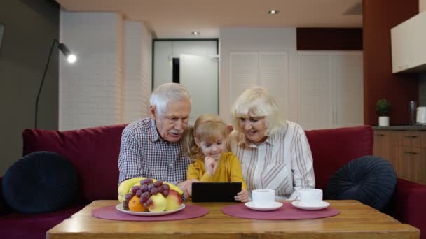 祖父母は小さな子供にタブレットを使って孫を教え、自宅でオンラインショッピングをしています — ストック動画