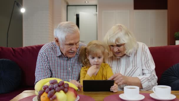 Dziadek i babcia siedzą w salonie i uczą małą wnuczkę za pomocą tabletu — Wideo stockowe