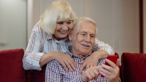 Szczęśliwa uśmiechnięta para seniorów z komórką w domu. Odpoczynek na kanapie w przytulnym salonie — Wideo stockowe