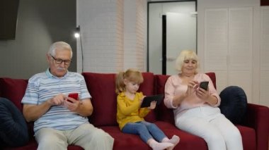 Büyükbaba ve torunlar evdeki kanepede oturup cep telefonu ve dijital tablet kullanıyorlar.