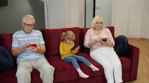 Abuelos y nieta se sientan en el sofá en casa y usan teléfonos móviles y tabletas digitales — Vídeos de Stock