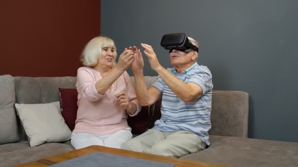 Starszy mężczyzna gra w wirtualnej rzeczywistości okulary słuchawkowe, kobieta śmieje się z nim akcja w domu — Wideo stockowe