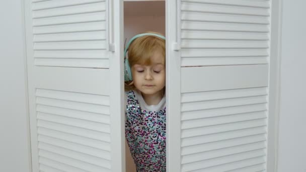 Niña pequeña con auriculares escuchando música y bailando graciosamente cerca del armario en casa — Vídeo de stock