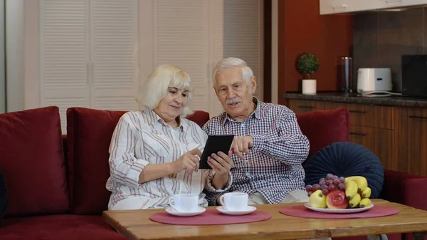 Ancianos abuelos de la vieja pareja hablando y usando tableta digital en casa. Compras por Internet — Foto de Stock