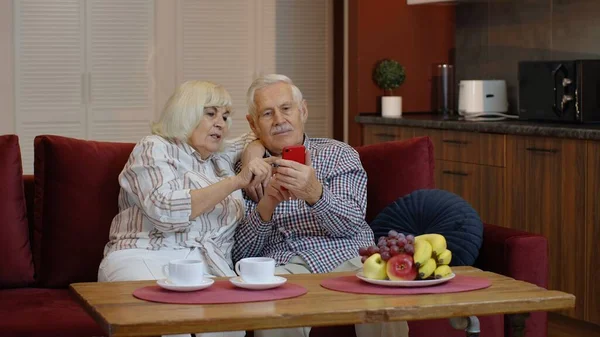 Senior old couple grandparents talking and using digital mobile phone at home. Online shopping