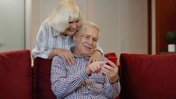 Ancianos abuelos de la vieja pareja hablando y usando la computadora del teléfono móvil en casa. Compras por Internet — Foto de Stock