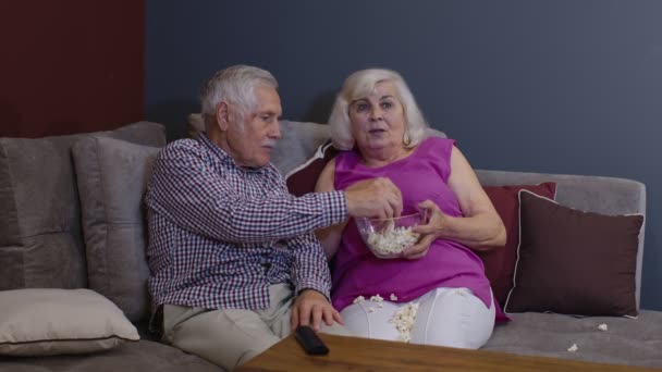 Налякана старша пара тримає попкорн, дивлячись фільм жахів, сидячи на дивані у вітальні — стокове відео