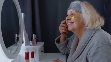 Yaşlı yaşlı bir kadın, cildiyle ilgileniyor. Gözlerinde kırışıklıklar var. Evde makyaj yapıyor.