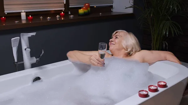 Senior woman grandmother is taking foamy bath, drinking champagne in luxury bathroom with candles — Stock Photo, Image