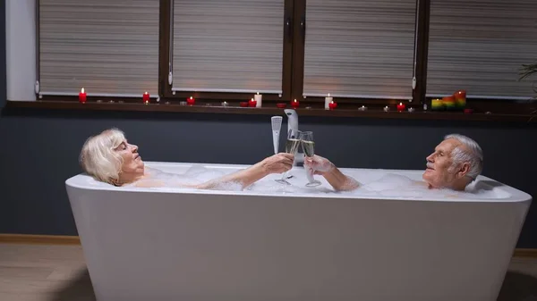 Senior couple in love lying in warm bath with bubbles, enjoying relaxation, drinking champagne — Stock Photo, Image