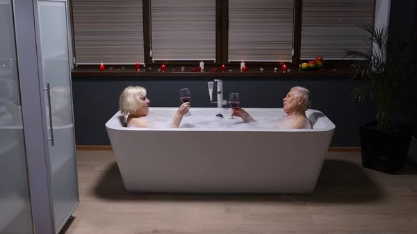 Senior couple grandfather and grandmother is taking foamy bath drinking red wine in luxury bathroom — Stock Photo, Image