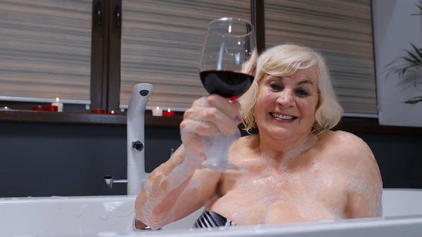 Ηλικιωμένη ηλικιωμένη γυναίκα ξαπλωμένη σε ζεστό μπάνιο με φυσαλίδες, απολαμβάνοντας χαλάρωση, πίνοντας κόκκινο κρασί — Φωτογραφία Αρχείου