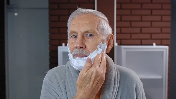Hombre mayor abuelo en albornoz afeitado con hoja de afeitar manual, mirando en el espejo. Punto de vista — Foto de Stock