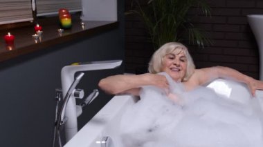 Seksi, yaşlı, yaşlı, sarışın kadın büyükannem lüks banyoda mumlarla köpüklü banyo yapıyor.