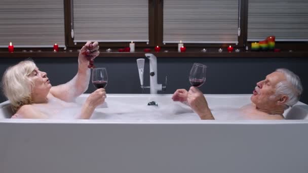 Kıdemli çift, sıcak bir küvette baloncuklarla uzanmış, rahatlamanın tadını çıkarıyor, kırmızı şarap içiyor. — Stok video