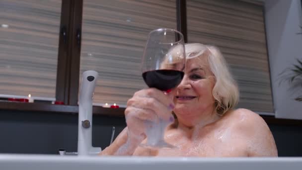 Mujer mayor abuela está tomando un baño espumoso, beber vino tinto en el baño de lujo con velas — Vídeo de stock