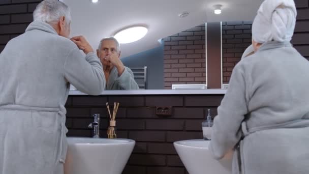 Senior couple grandmother and grandfather brushing teeth and looking into a mirror at bathroom — Stock Video
