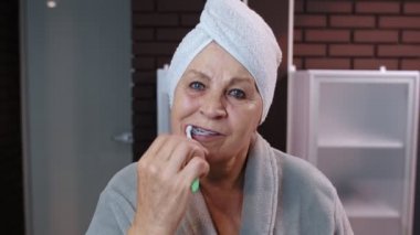 Havluyla dişlerini fırçalayan ve duştan sonra aynaya bakan yaşlı bir kadın.