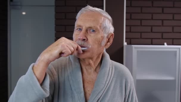 Senior man grandfather in bathrobe brushing teeth, looking into mirror. Morning hygiene at bathroom — Stock Video