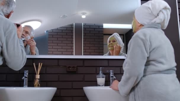 Oudere stel man en vrouw doen 's ochtends hygiëneprocedures en kijken in een spiegel — Stockvideo