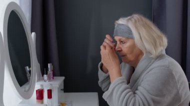 Yaşlı, yaşlı bir kadın, kırışıklık önleyici göz bandı takıyor, evde makyaj yapıyor.