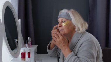 Yaşlı, yaşlı bir kadın, kırışıklık önleyici göz bandı takıyor, evde makyaj yapıyor.